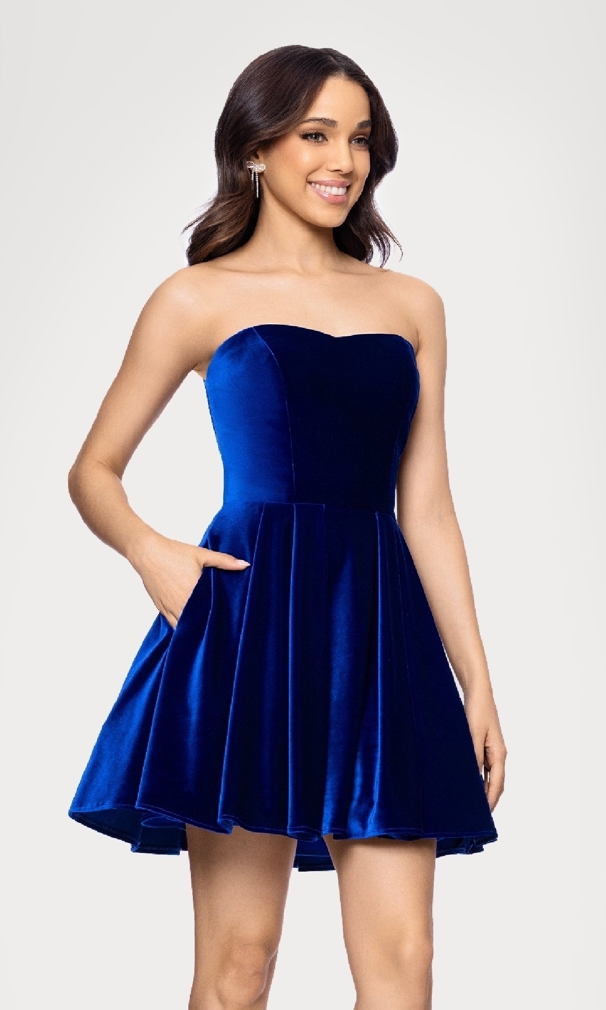 A woman in the Blondie Nites 5718BN Short Velvet Hoco Dress—a strapless, royal blue, flared cocktail dress with pockets—smiles against a plain light background.